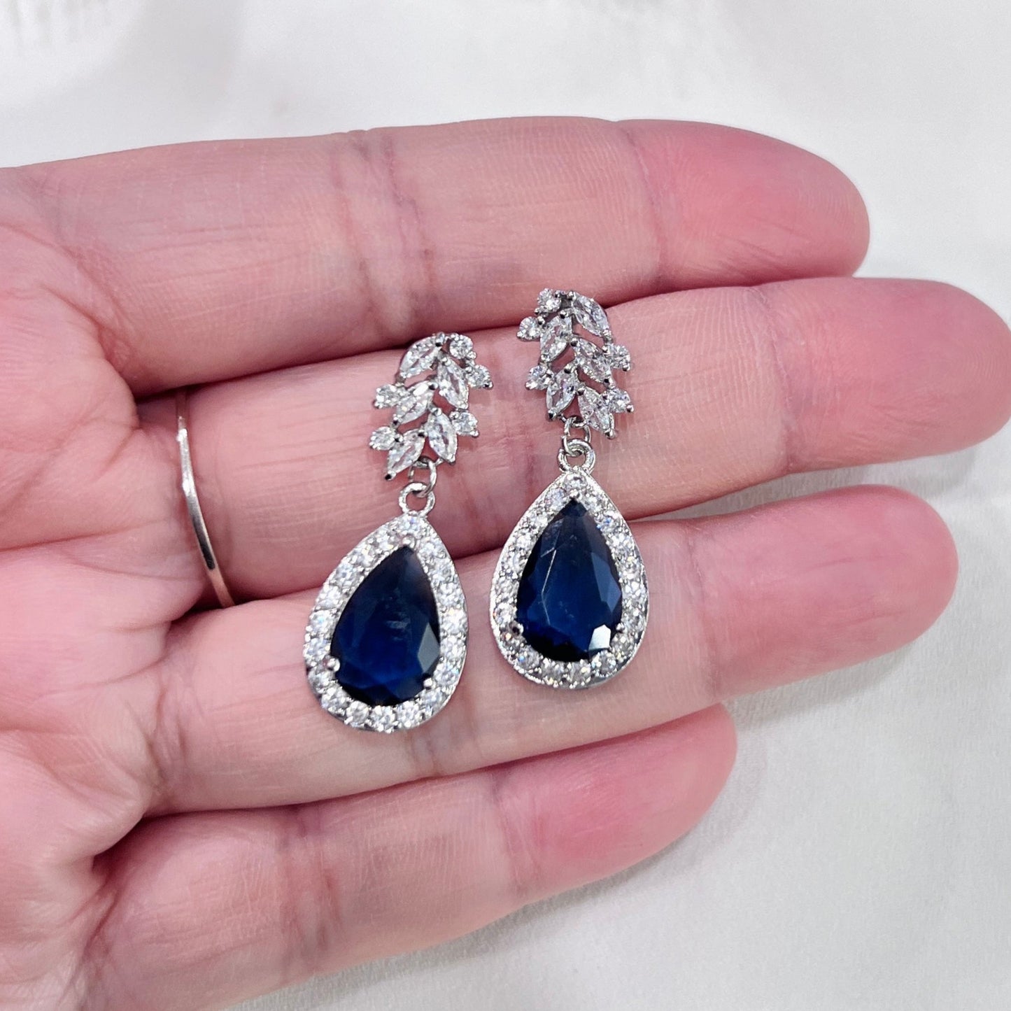 Teardrop-shaped sapphire blue gemstone earrings with silver detailing held in a hand.