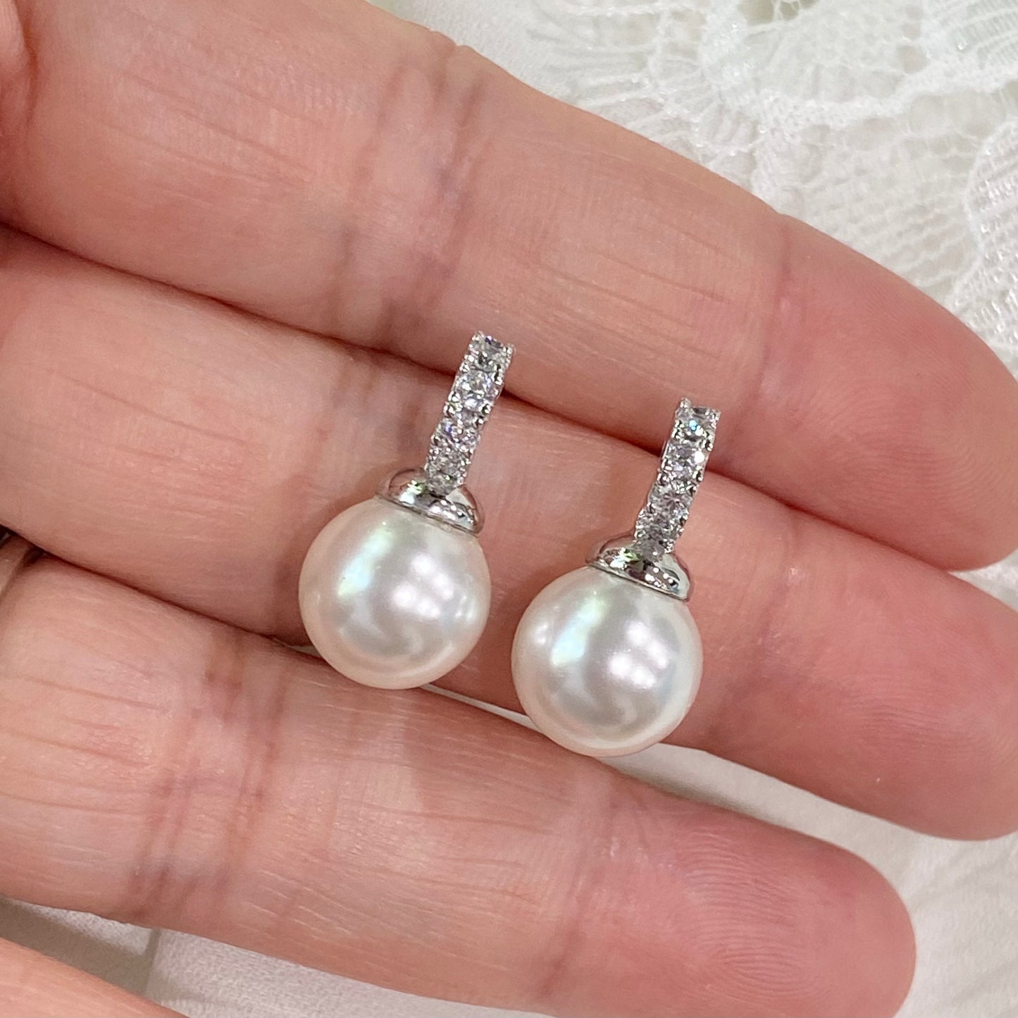 Bridal white pearl drop earrings with crystal accents photographed on soft lace fabric background