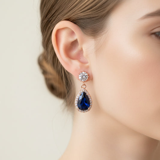 Pair of blue teardrop earrings with rose gold and clear gemstones held in a hand against a neutral background.
