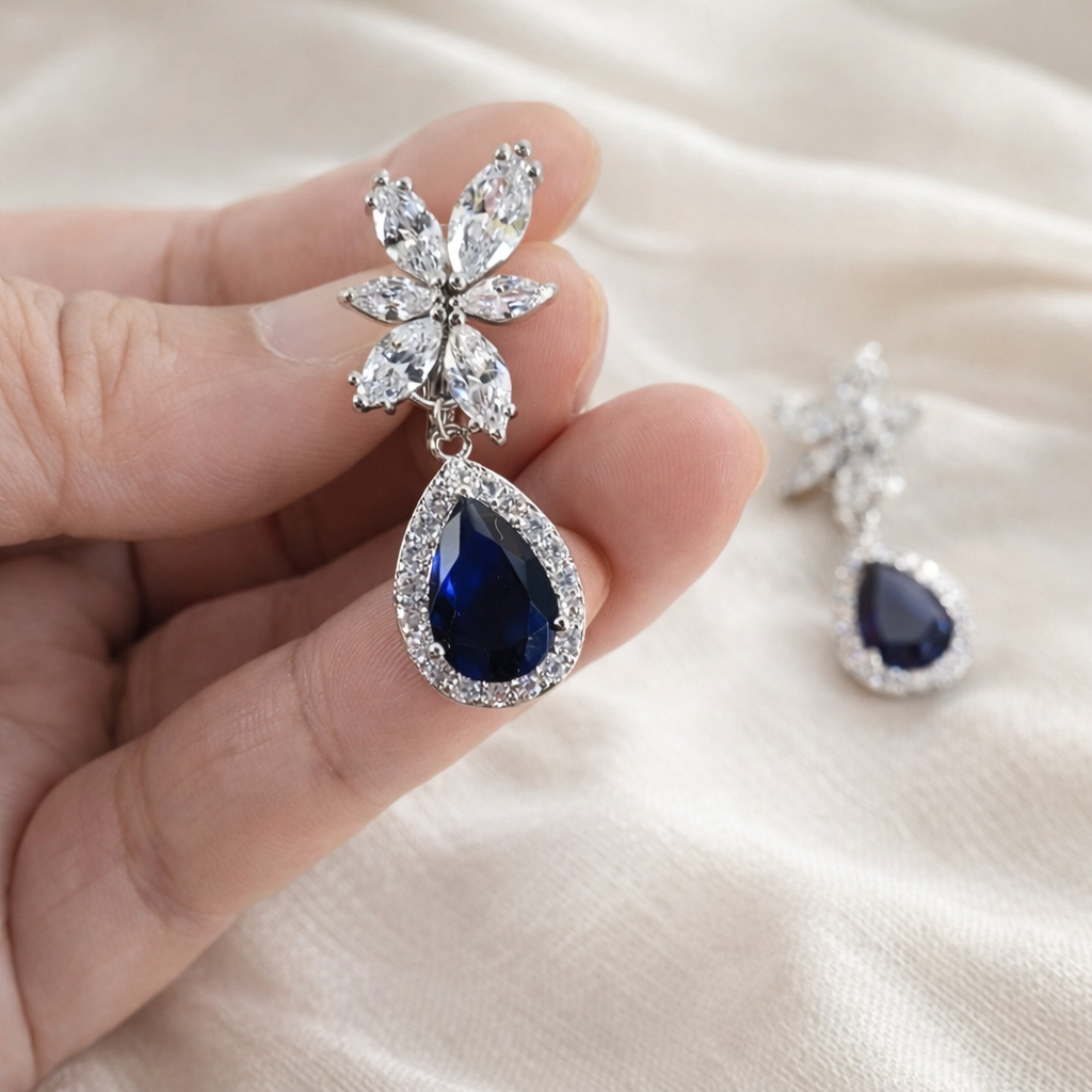 Earring with blue teardrop gem and clear stones held by a hand on a soft fabric background