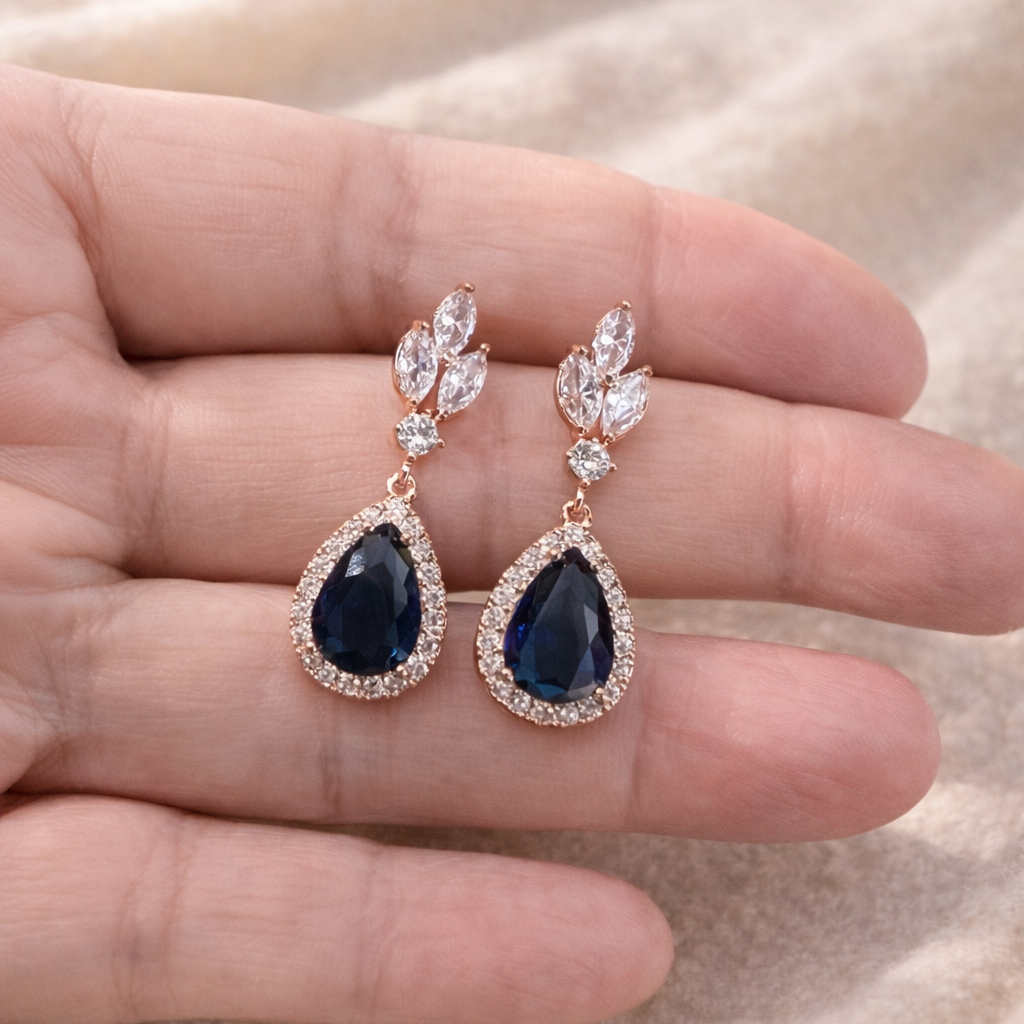 Pair of blue teardrop earrings held between fingers on a beige fabric background