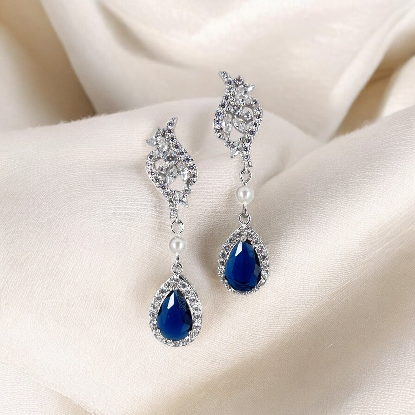 Pair of blue teardrop earrings with silver and pearl details on a beige fabric background