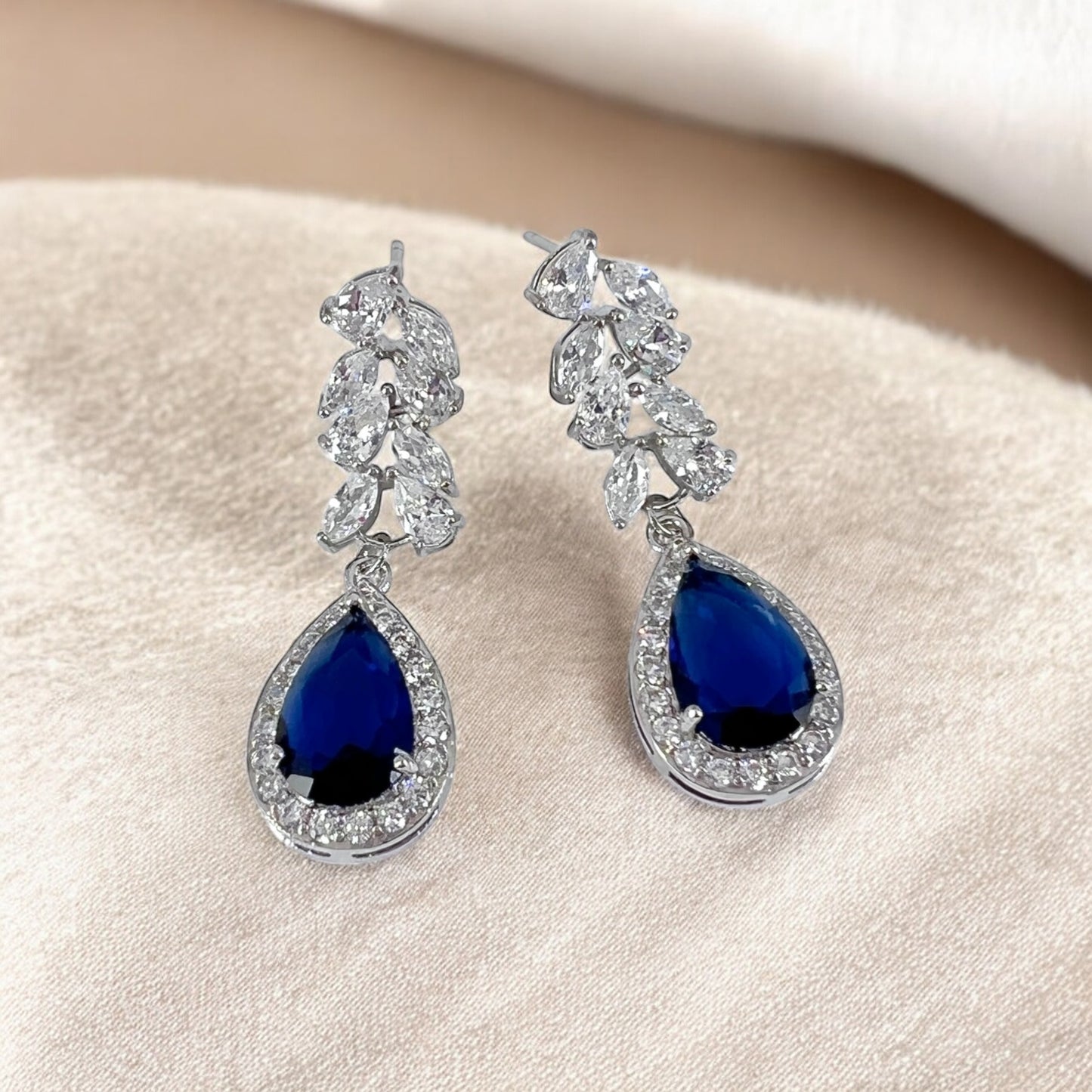 Sapphire blue teardrop bridal earrings with crystal halo photographed on soft neutral fabric