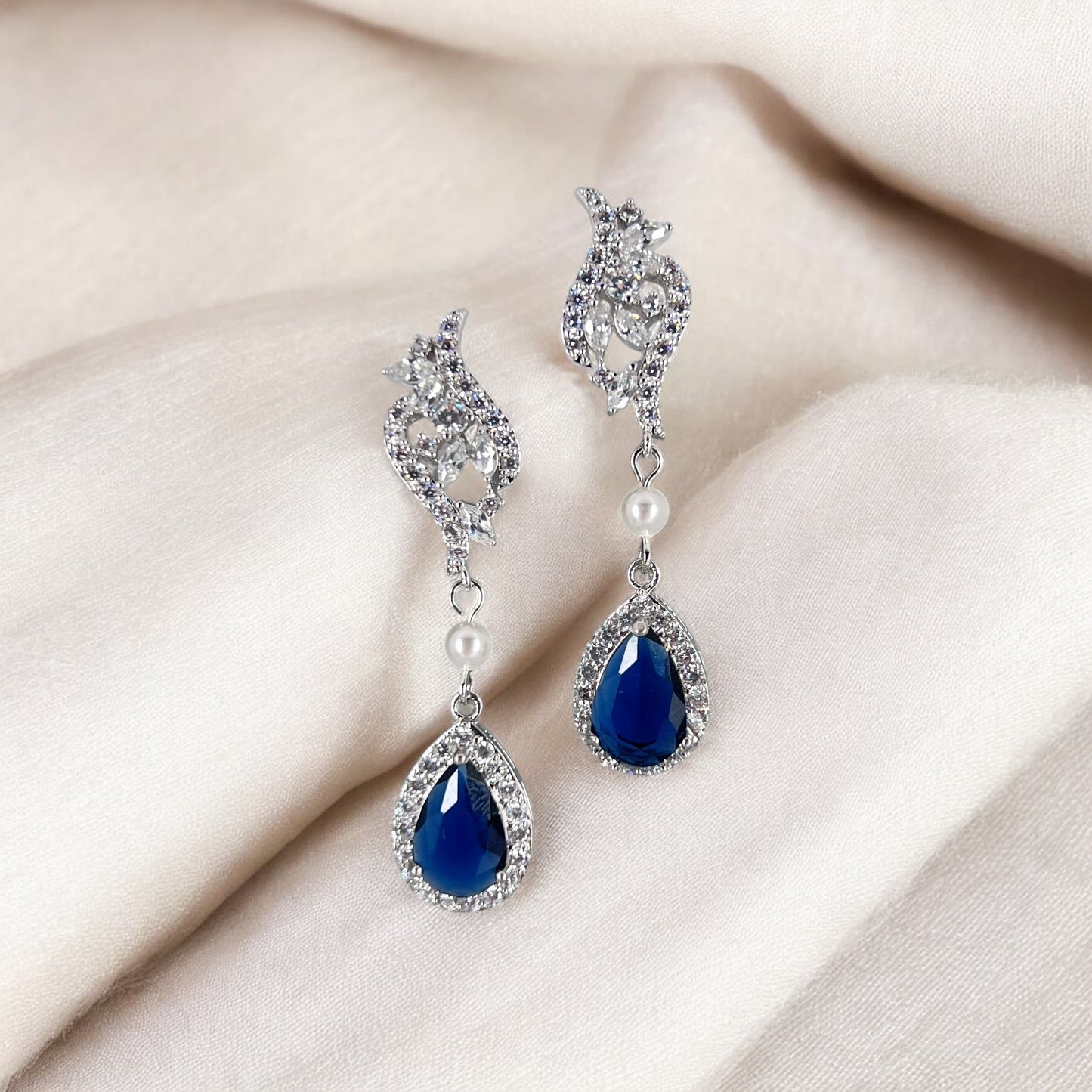Pair of blue teardrop earrings with silver and pearl details on a beige fabric background