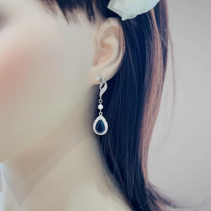 Close-up of an earring with a blue gemstone worn by a person with dark hair and a white flower in their hair.