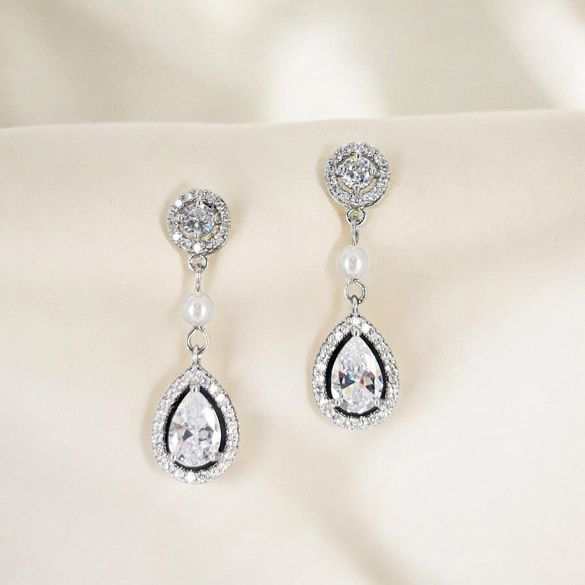 Clear crystal teardrop bridal earrings with pearl connector photographed on soft neutral fabric