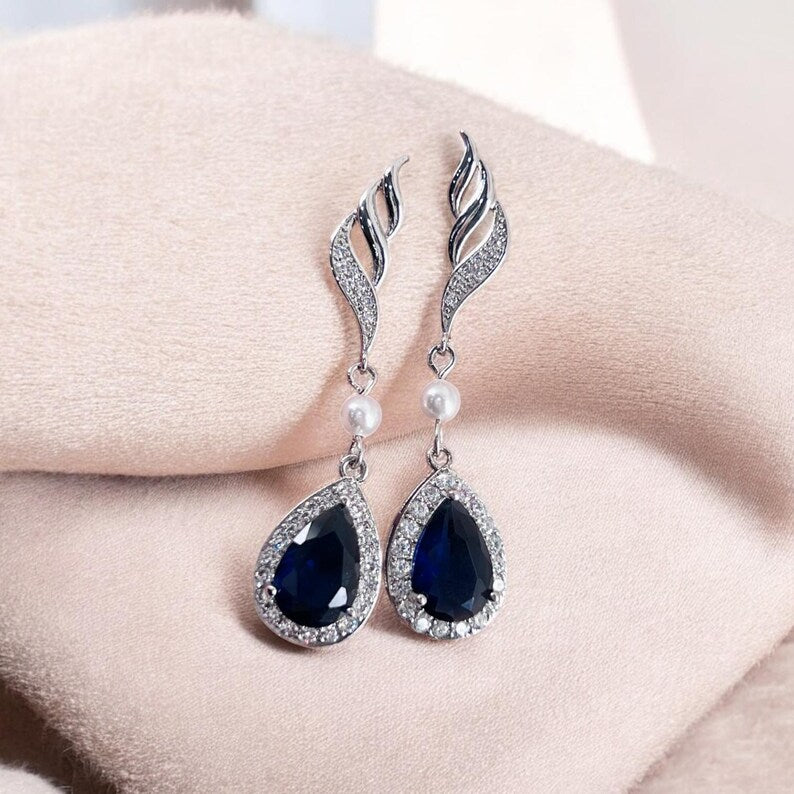 Pair of blue teardrop earrings with silver and pearl accents on a soft pink fabric background.